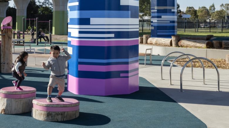 Children enjoy repurposed logs in playground beneath elevated rail.