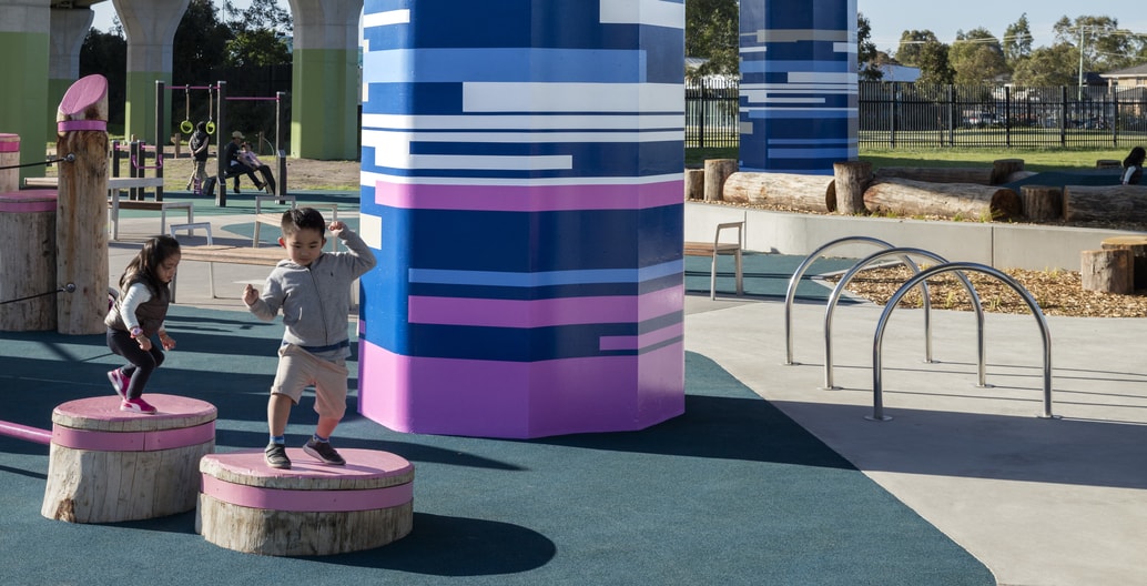 Children enjoy repurposed logs in playground beneath elevated rail.